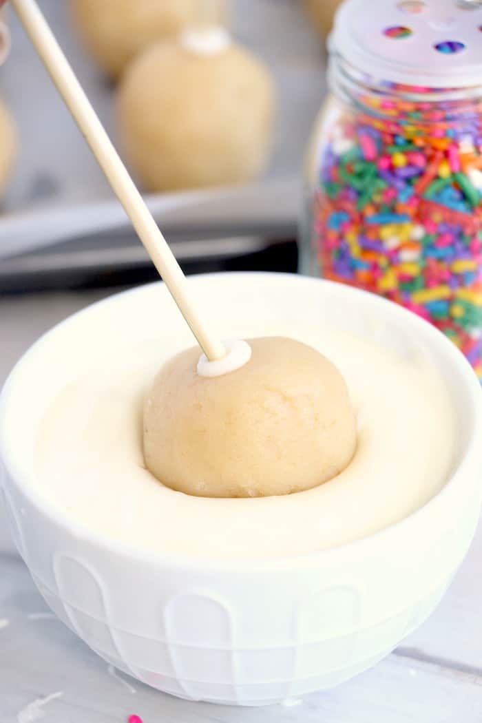 A cake pop on a wooden skewer being dipped into melted white chocolate.