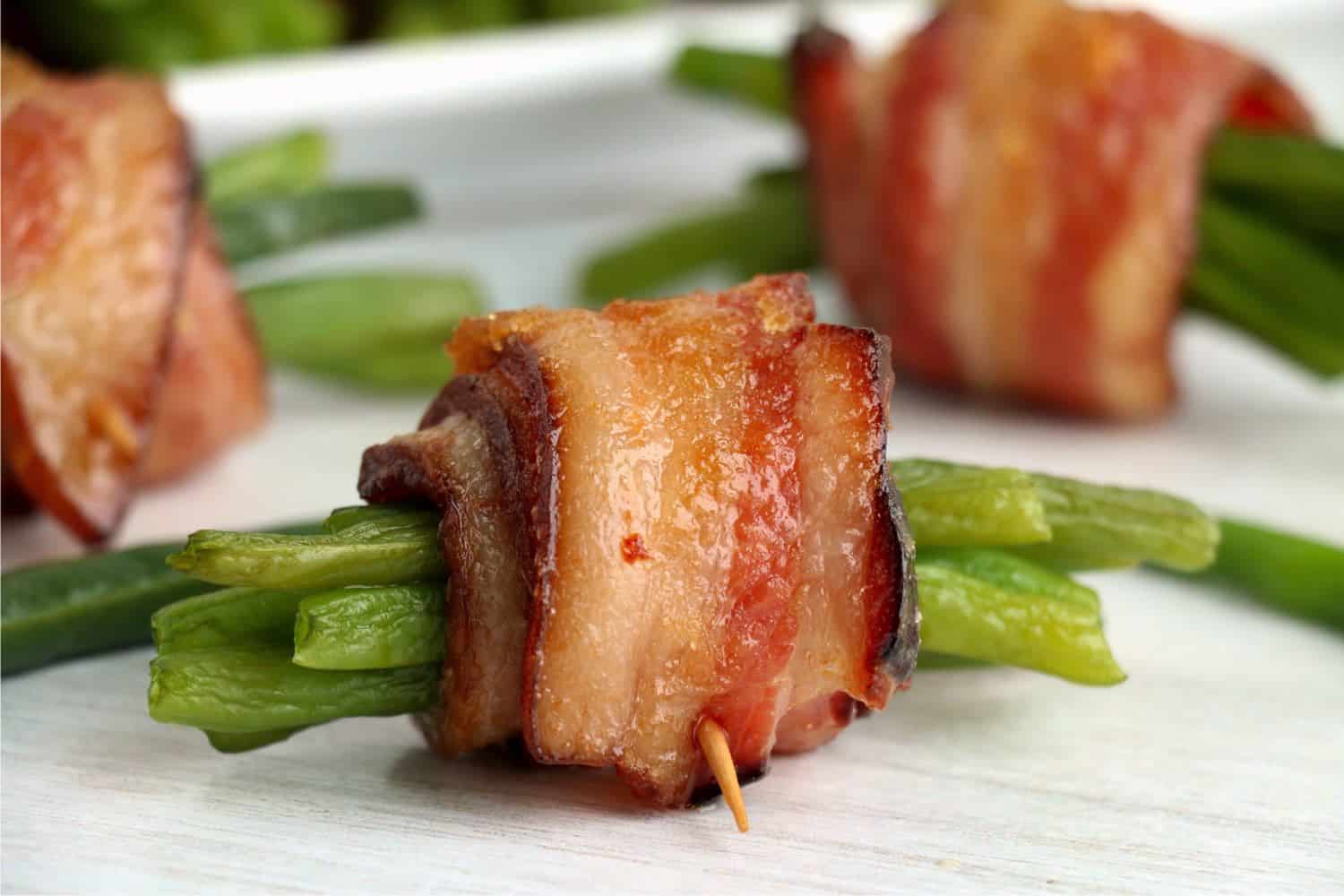 Cooked bacon wrapped green beans. One bundle is close up and two more are blurry in the background