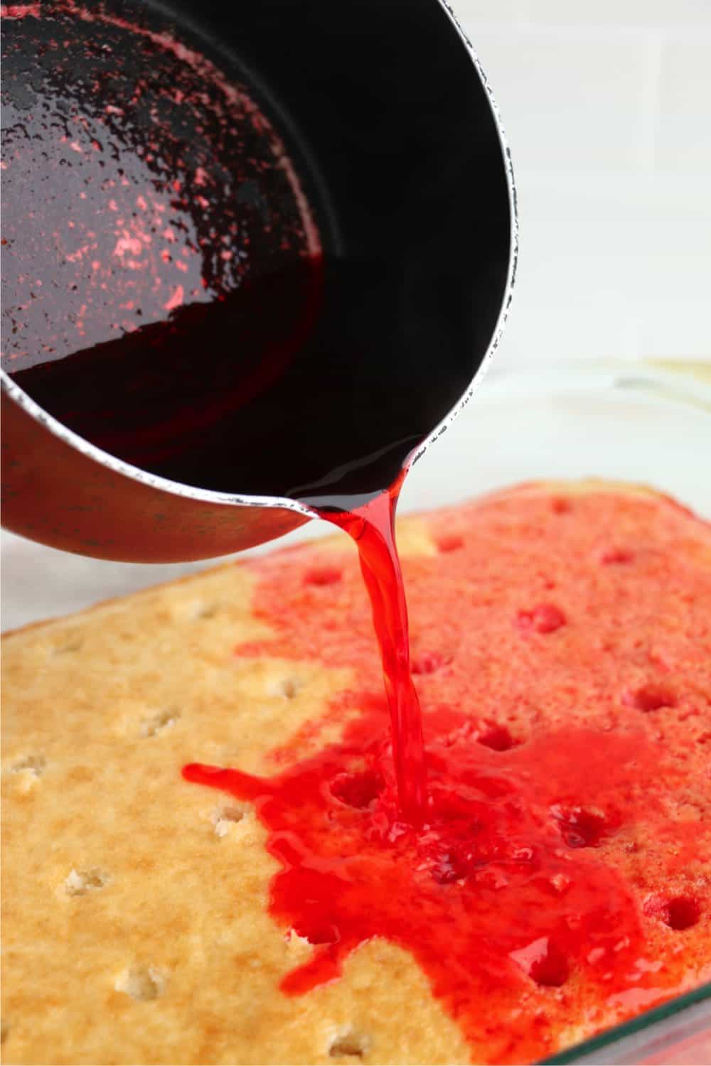 Raspberry jello being poured into a baked white cake with holes poked throughout.