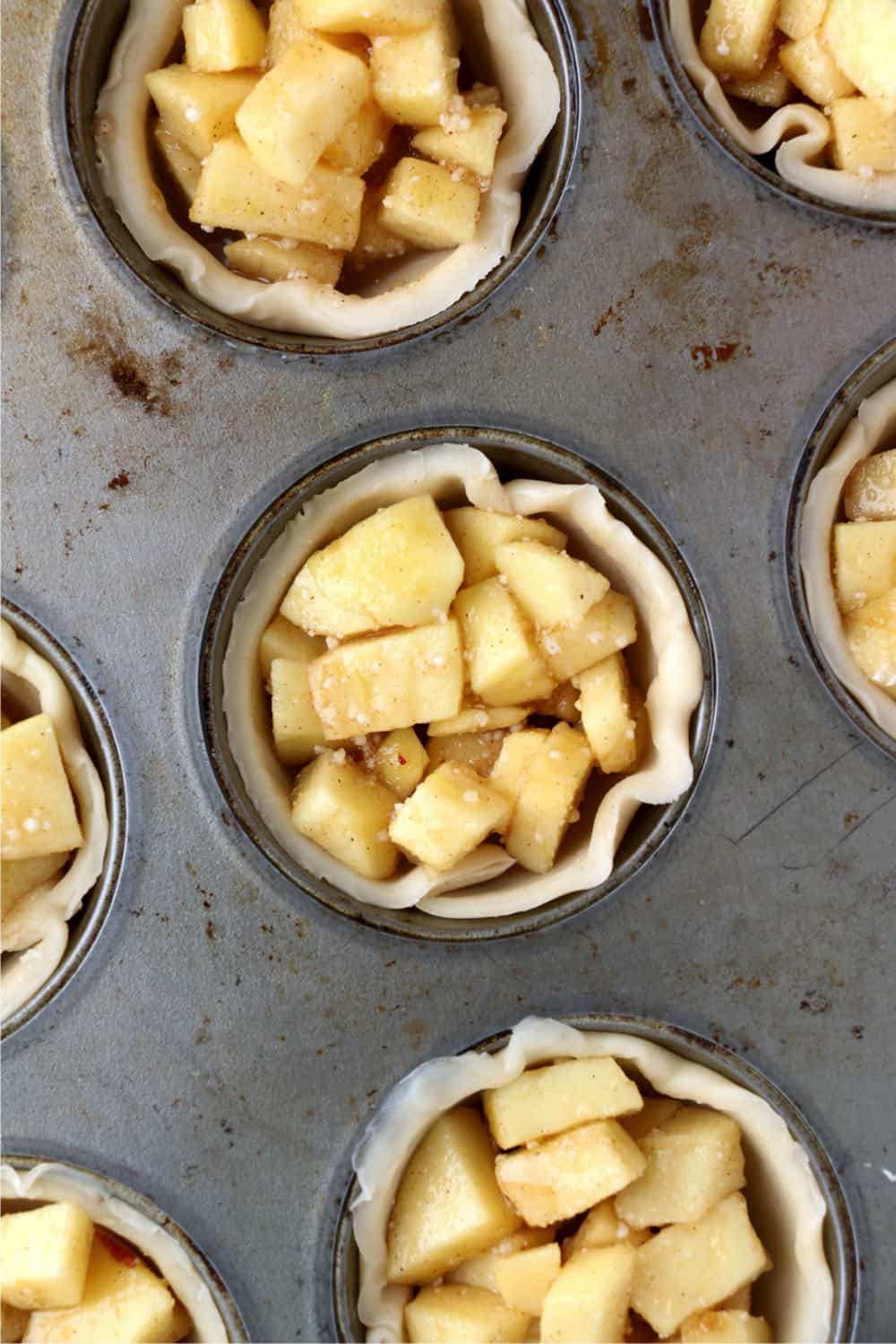 Miniature pie crusts in a muffin tin, filled with apple filling