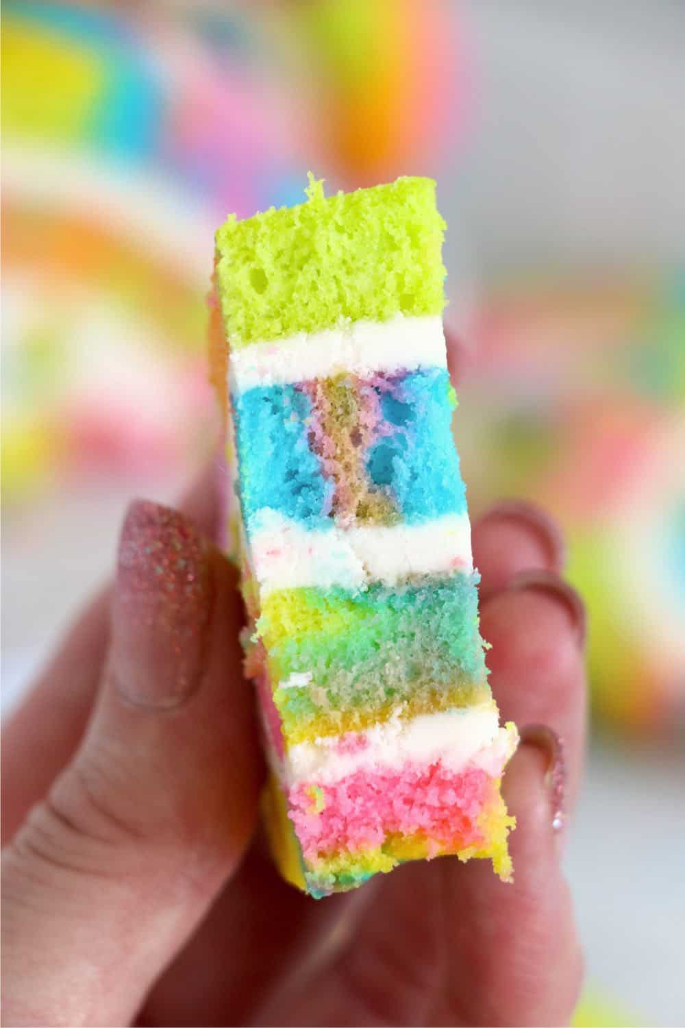 A hand with polished anils holding a slice of rainbow cake with layers of buttercream frosting