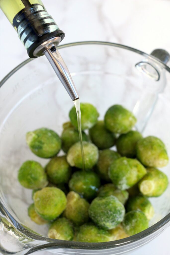 Closeup shot of oil being poured over frozen brussel sprouts in bowl.