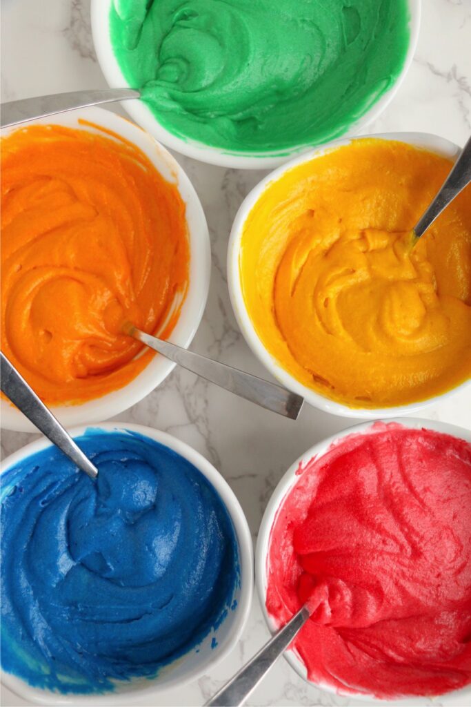 Overhead shot of green, orange, yellow, blue, and red frosting in separate bowls