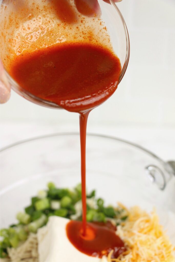 Closeup shot of Buffalo sauce being poured into bowl filled with chicken salad ingredients