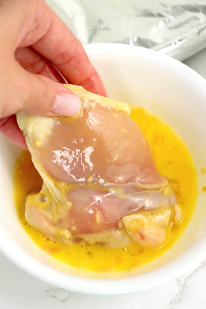Hand dipping a chicken thigh into a bowl of egg mixture