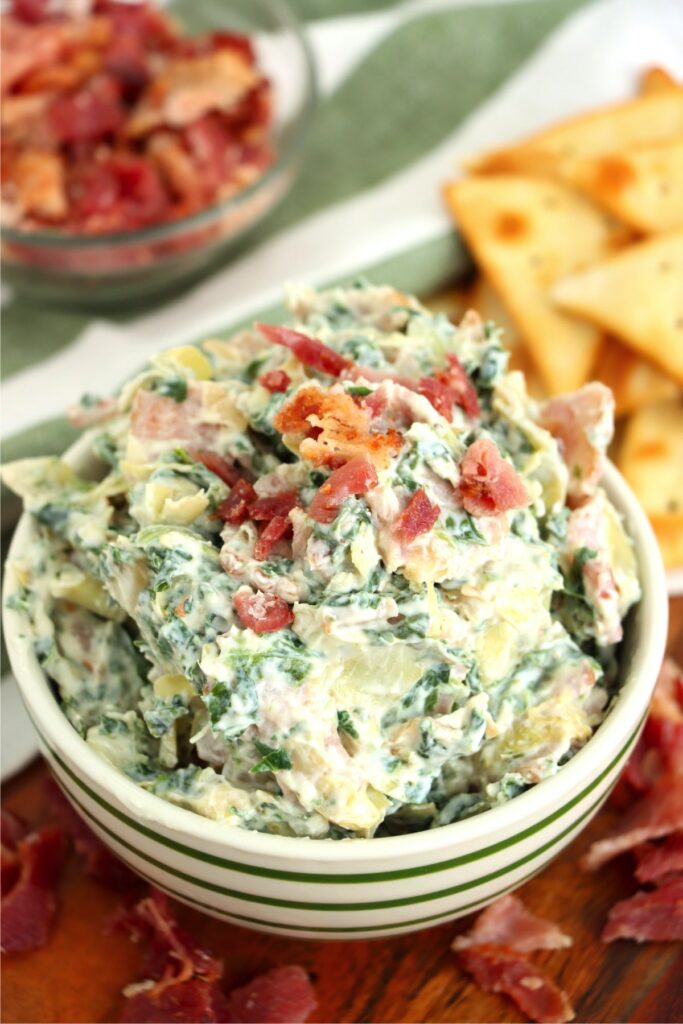 Closeup shot of bowlful of spinach artichoke dip with bacon next to pita chips