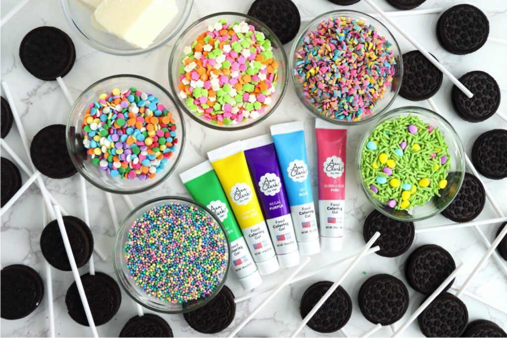 Overhead shot of sprinkles and almond bark in bowls, surrounding tubes of food coloring gel with Oreos on sticks all around