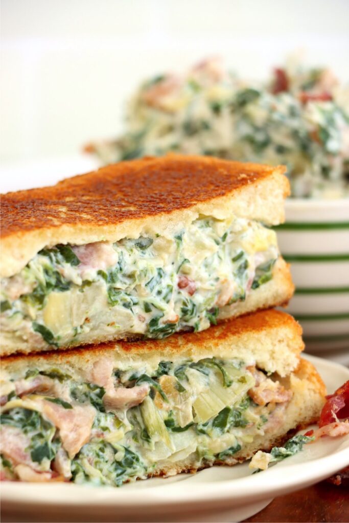 Closeup shot of tow spinach artichoke grilled cheese sandwich halves stacked atop one another on plate.
