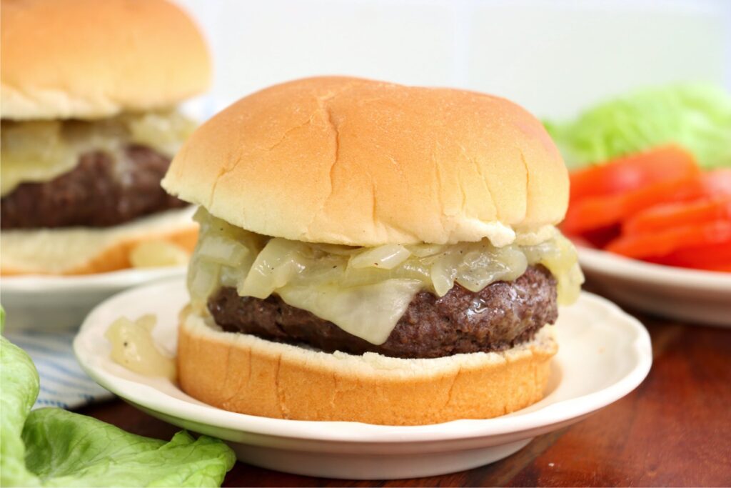 Closeup shot of grilled onion cheddar burger on plate with another burger and lettuce and tomato on plates in background