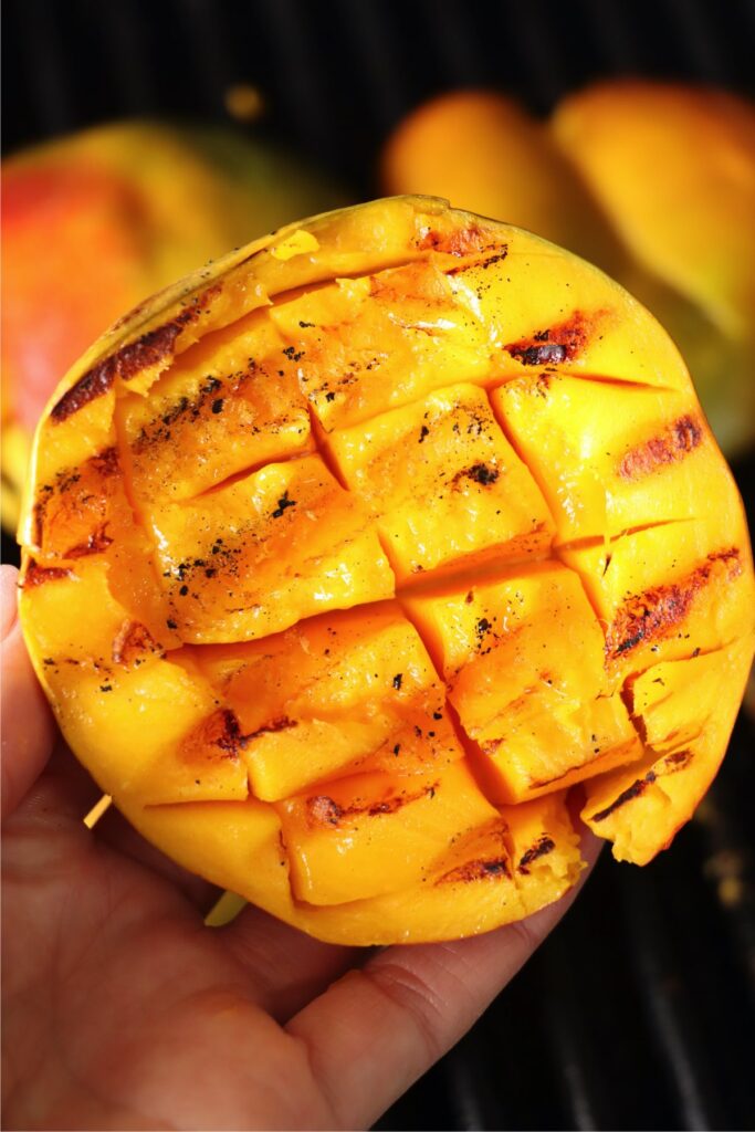 Closeup shot of hand holding grilled mango over grill grate with more grilled mangoes