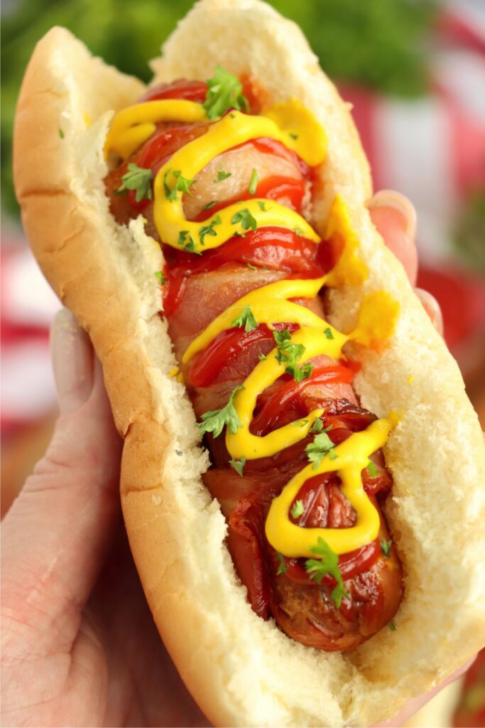 Closeup shot of hand holding air fryer bacon wrapped hot dog in bun topped with ketchup and mustard. 