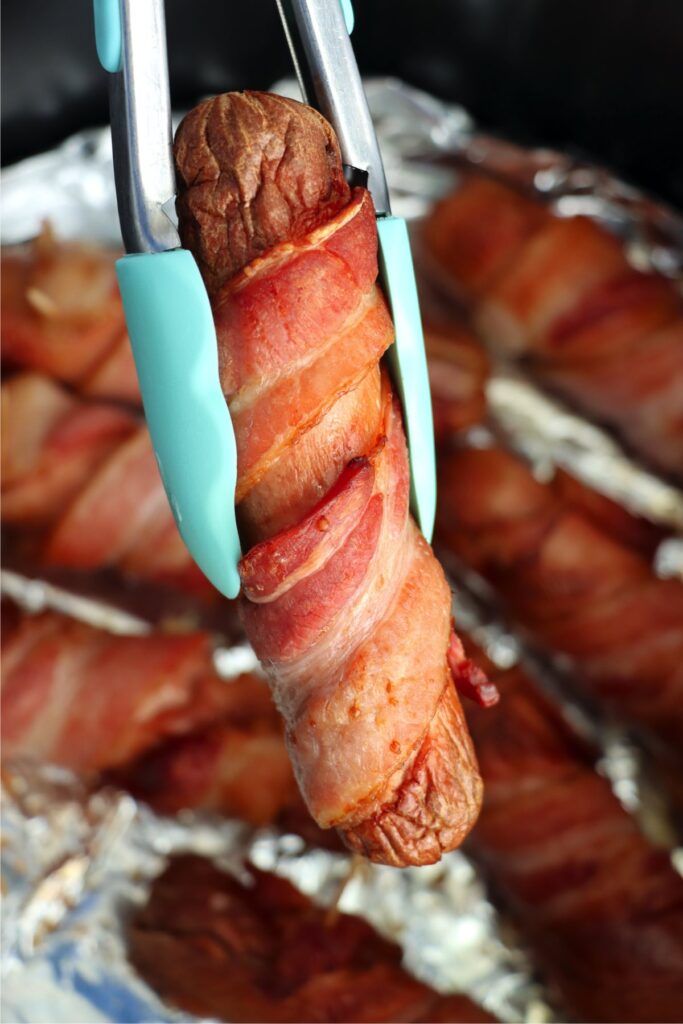 Closeup shot of tongs holding a bacon wrapped hot dog with more bacon wrapped hot dogs in the background.