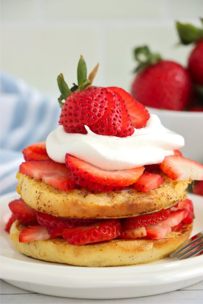 Closeup shot of English muffin French toast topped with strawberries and whipped cream on plate.