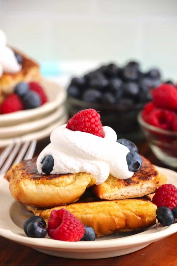 Closeup shot of stack of hot dog bun French toast topped with whipped cream and berries on plate. 