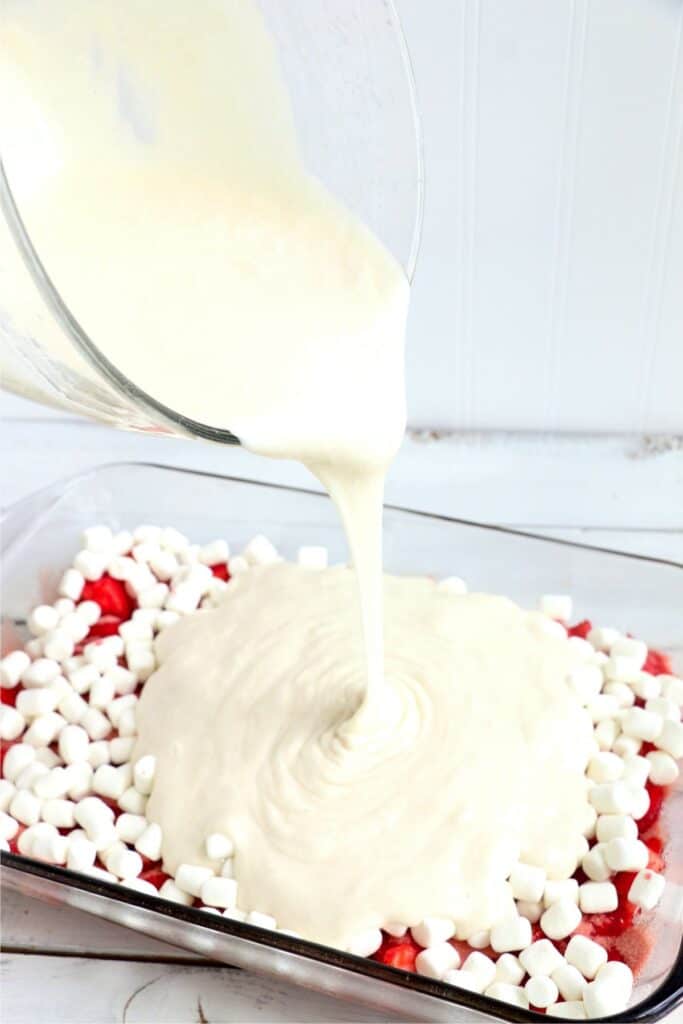 Cake batter being poured into baking dish with strawberries, Jello, and min marshmallows. 