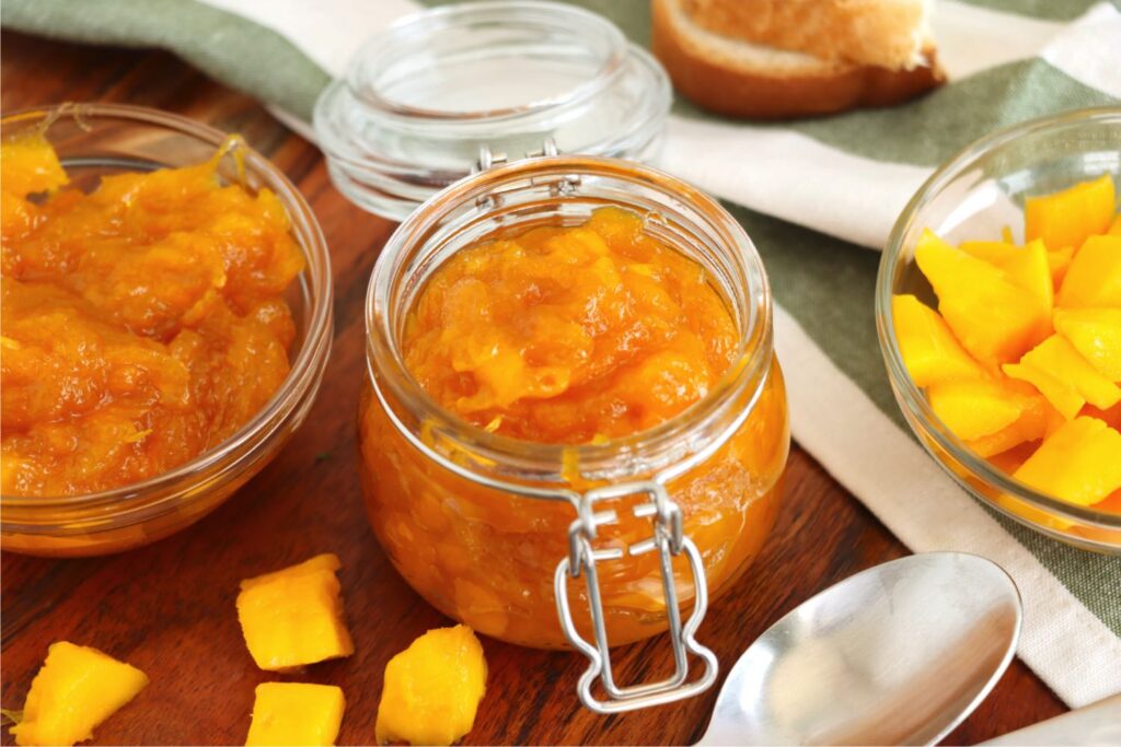 Jarful of homemade mango jam next to bowl of jam and bowl of cubed mango