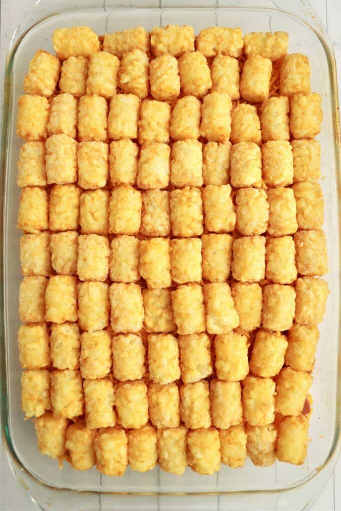 Overhead shot of tater tots placed on top of cheese, chili, and hot dog layers in baking dish