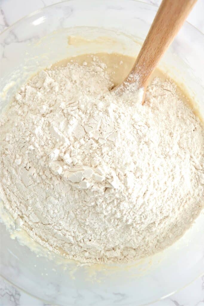 Overhead shot of spoon in bowlful of wet dough mixture topped with flour.