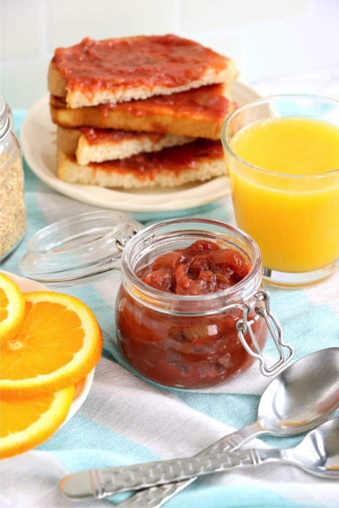 Glass jar full of homemade rhubarb jam with bread topped with rhubarb jam on plate in background