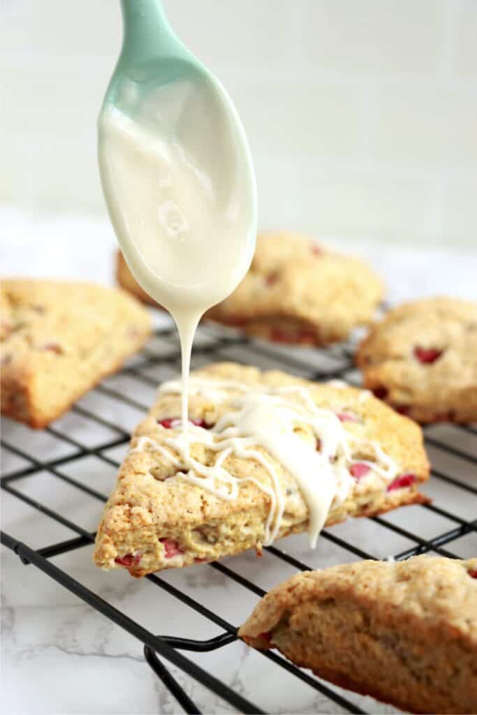 Closeup shot of vanilla glaze being drizzled over homemade rhubarb scones with spoon. 