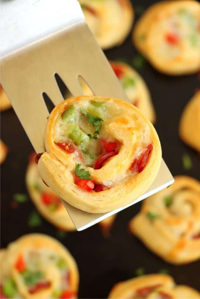 Closeup overhead shot of golden brown pizza pinwheel on spatula over baking sheet with more pinwheels