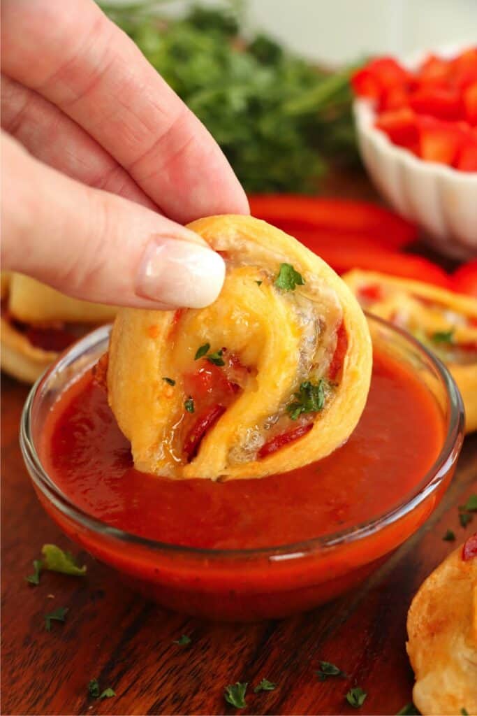 Hand dipping a pizza pinwheel into pizza sauce in a small bowl. 