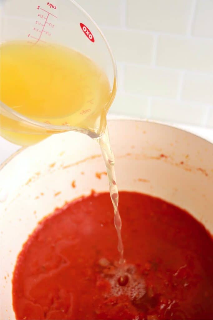 Chicken broth being poured into skillet with vegetables and crushed tomatoes