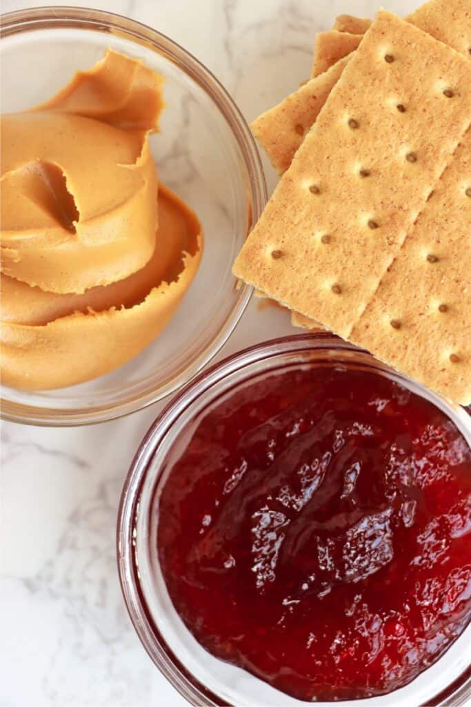 Overhead shot of graham crackers next to a bowl of peanut butter and another bowl of jelly. 