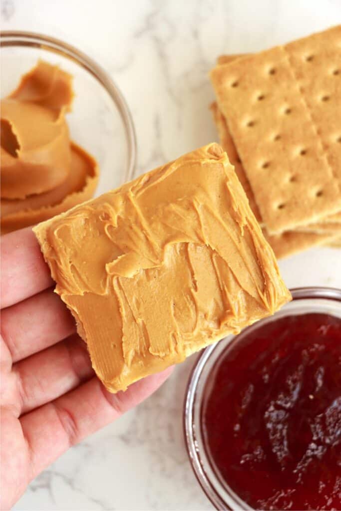 Overhead shot of hand holding a graham cracker square topped with peanut butter. 