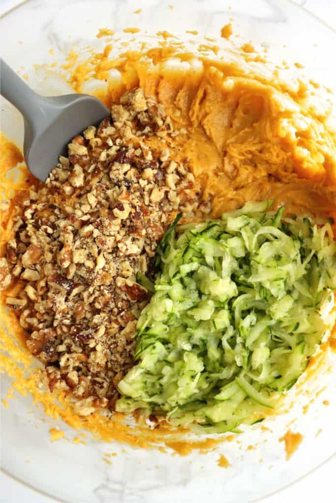 Overhead shot of grated zucchini and chopped walnuts on top of muffin batter in mixing bowl. 