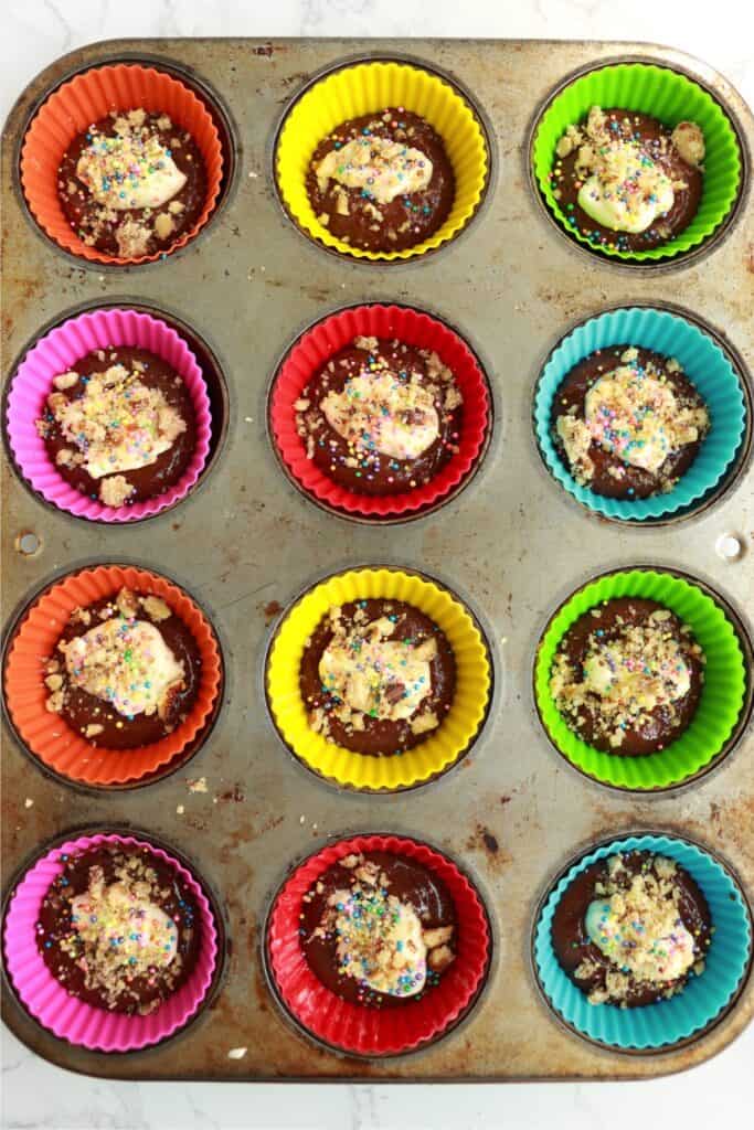 Cupake liners half filled with chcolate cake batter topped with cream cheese mixture, sprinkles, and nuts.