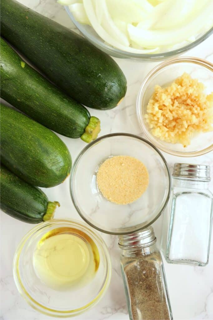 Overhead shot of whole zucchini along with the rest of the sauteed zucchini and onion ingredients in individual bowls on table