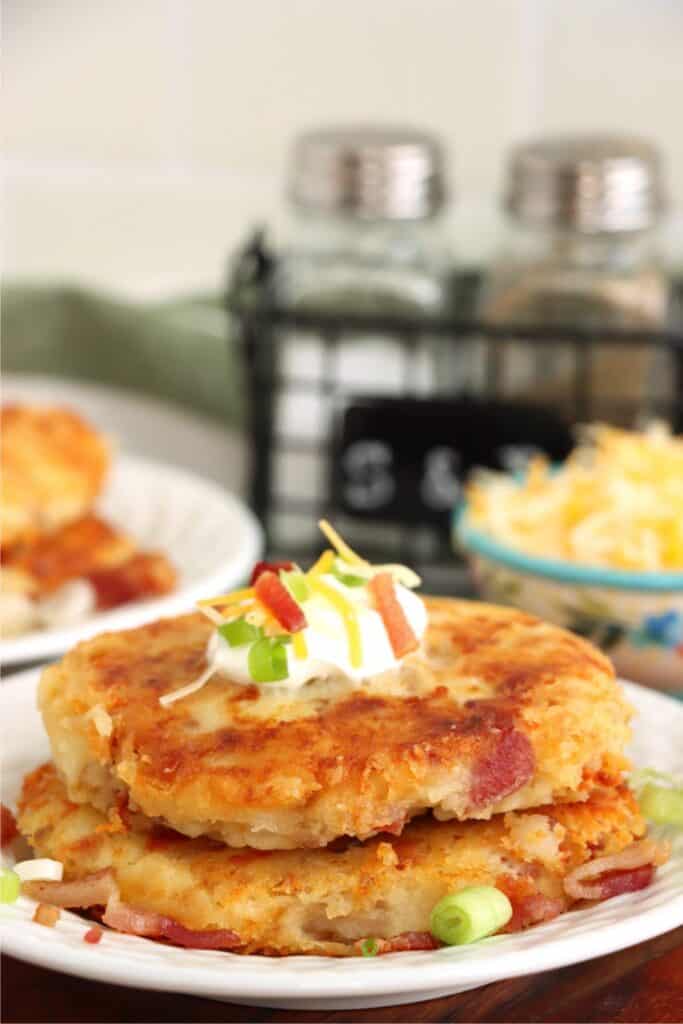 Closeup shot of two bacon and cheese leftover mashed potato patties stacked atop one another on plate and topped with sour cream and green onions. 