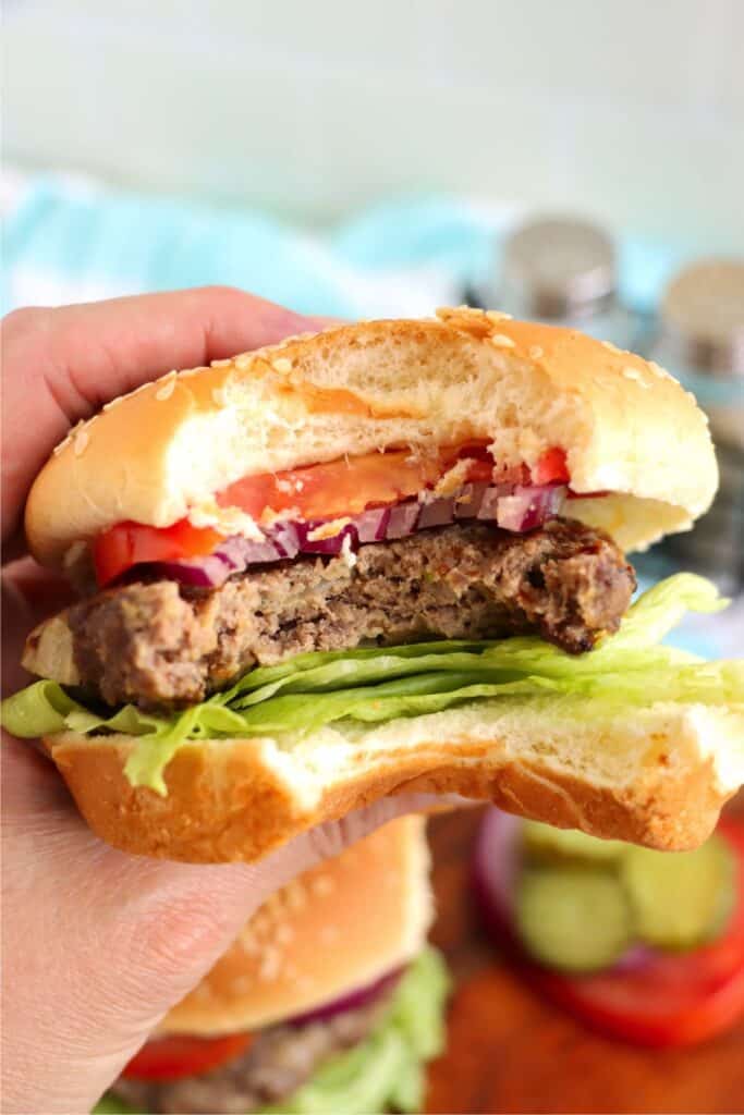 Hand holding a classic American hamburger topped with lettuce, tomato, onion, and pickles with bite taken out. 