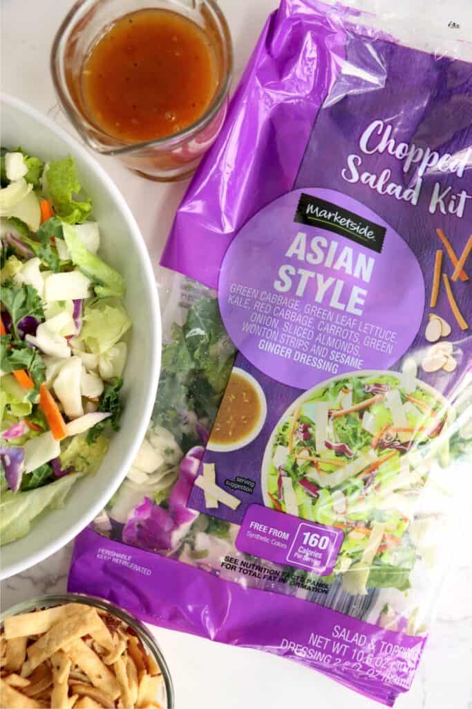 Overhead shot of bowlful of Asian salad next to cup of dressing and bag of Asian salad. 
