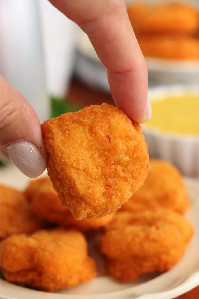Closeup shot of hand holding a Tyson air fryer chicken nugget in front of a plateful of more chicken nuggets. 