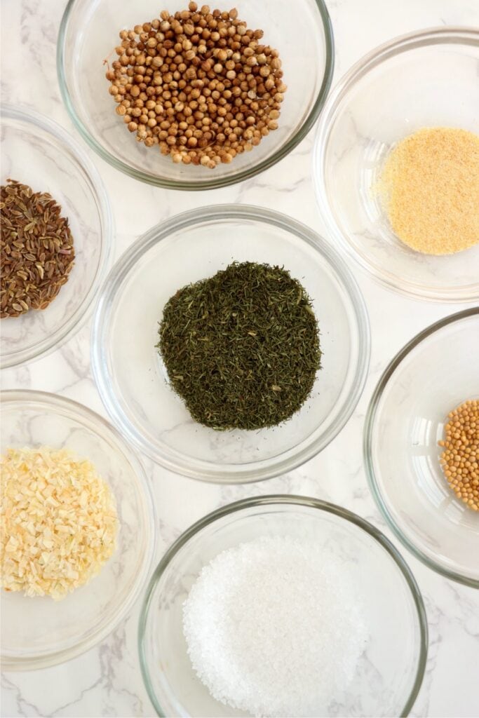 Overhead shot of individual dill pickle seasoning ingredients in bowls on table. 