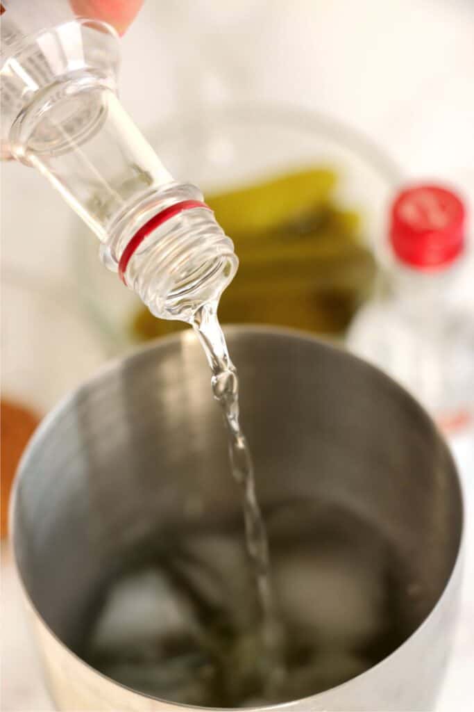 Closeup shot of vodka being poured into cocktail shaker filled with ice and pickle juice.