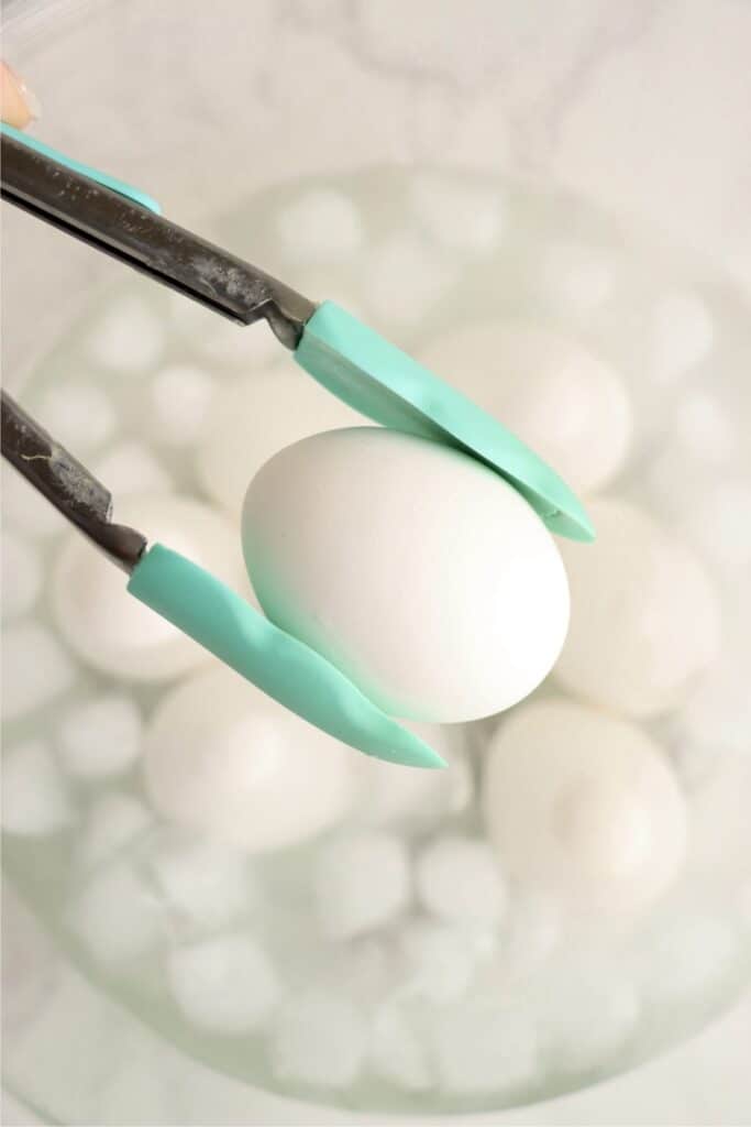 Closeup overhead shot of tongs holding an air fryer soft boiled egg over bowl filled with eggs in an ice water bath.