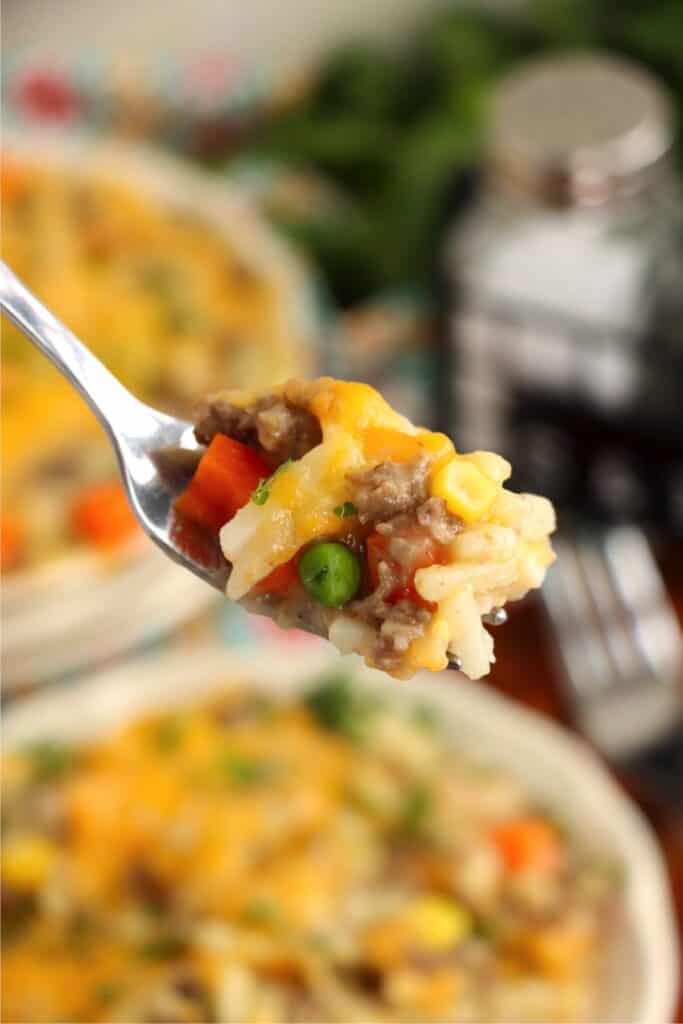 Closeup shot of forkful of ground beef and hash brown casserole over plate of more casserole. 