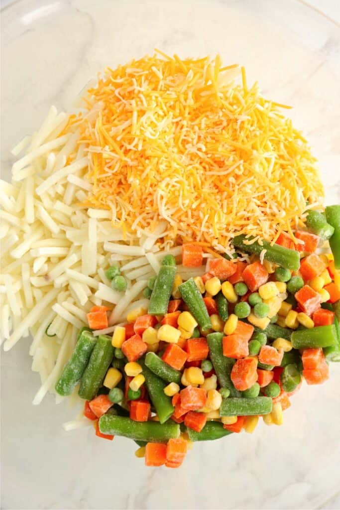 Closeup overhead shot of bowlful of frozen veggies, hash browns, and shredded cheese. 