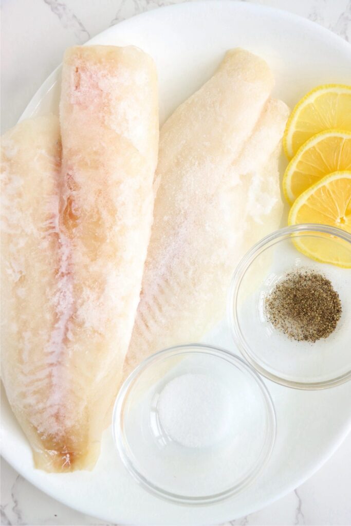 Overhead shot of frozen cod fillets, salt, pepper, and lemon slices on plate. 