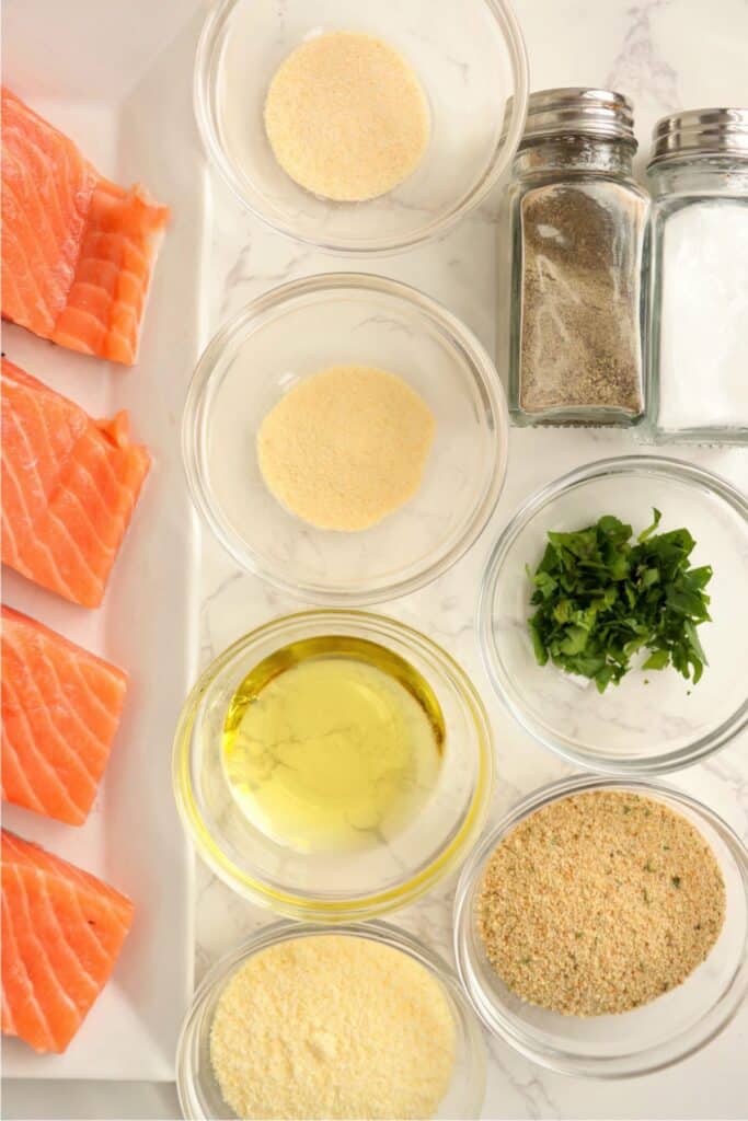 Overhead shot of salmon fillets on plate next to bowls of seasoning, breadcrumbs, and oil. 