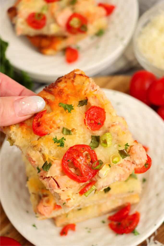 Closeup shot of hand holding a slice of salmon pizza overplateful of more pizza.