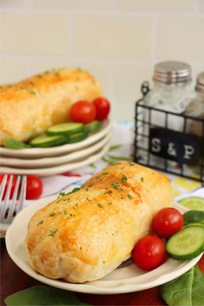 Plate with salmon Wellington, cherry tomatoes, and cucumber slices with another plate of salmon Wellington in background. 
