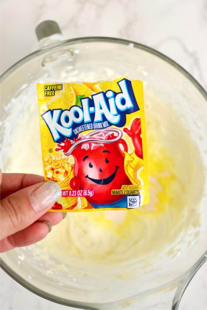 Overhead shot of packet of lemonaid Kool-Aid being held over bowlful of mixed cream cheese ingredients.